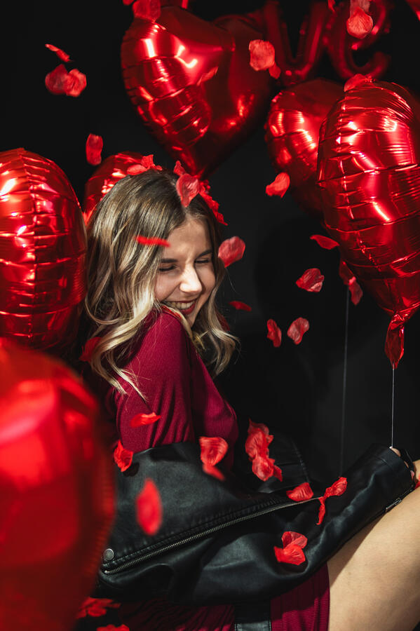 Portret kobiety wśród czerwonych balonów i płatków róż podczas walentynkowej sesji.