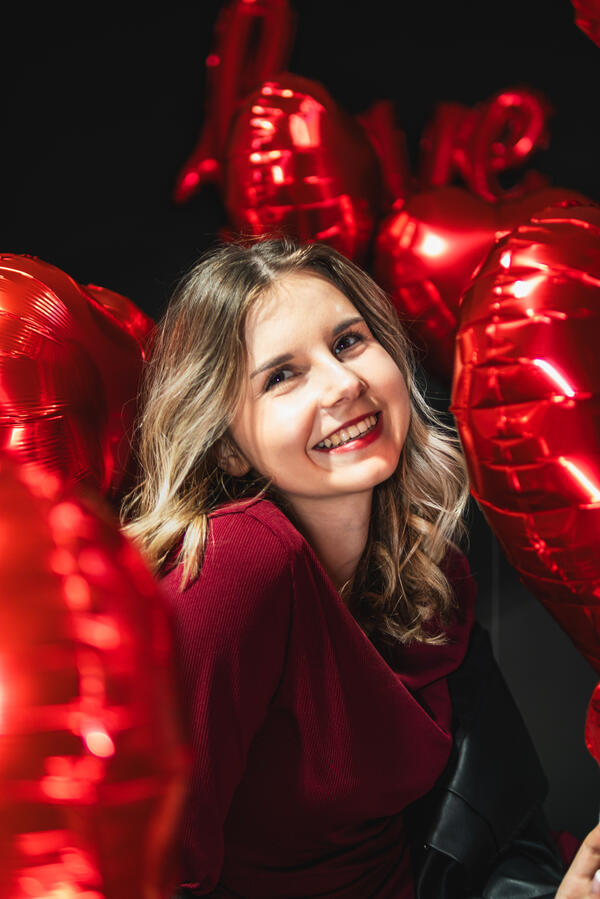 Uśmiechnięta kobieta na tle czerwonych balonów w kształcie serc podczas sesji portretowej.