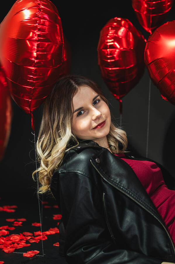 Portret kobiety w czerwonej sukience i czarnej kurtce wśród czerwonych balonów w kształcie serc.